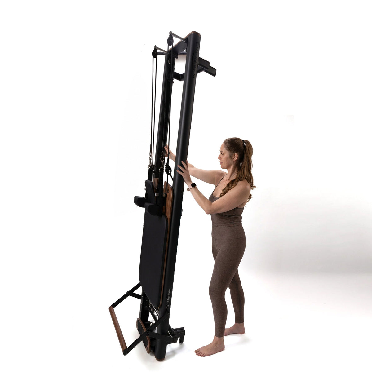 Woman standing beside upright black Peak Pilates fit Reformer with Walnut accents, showcasing folded footbar, retracted