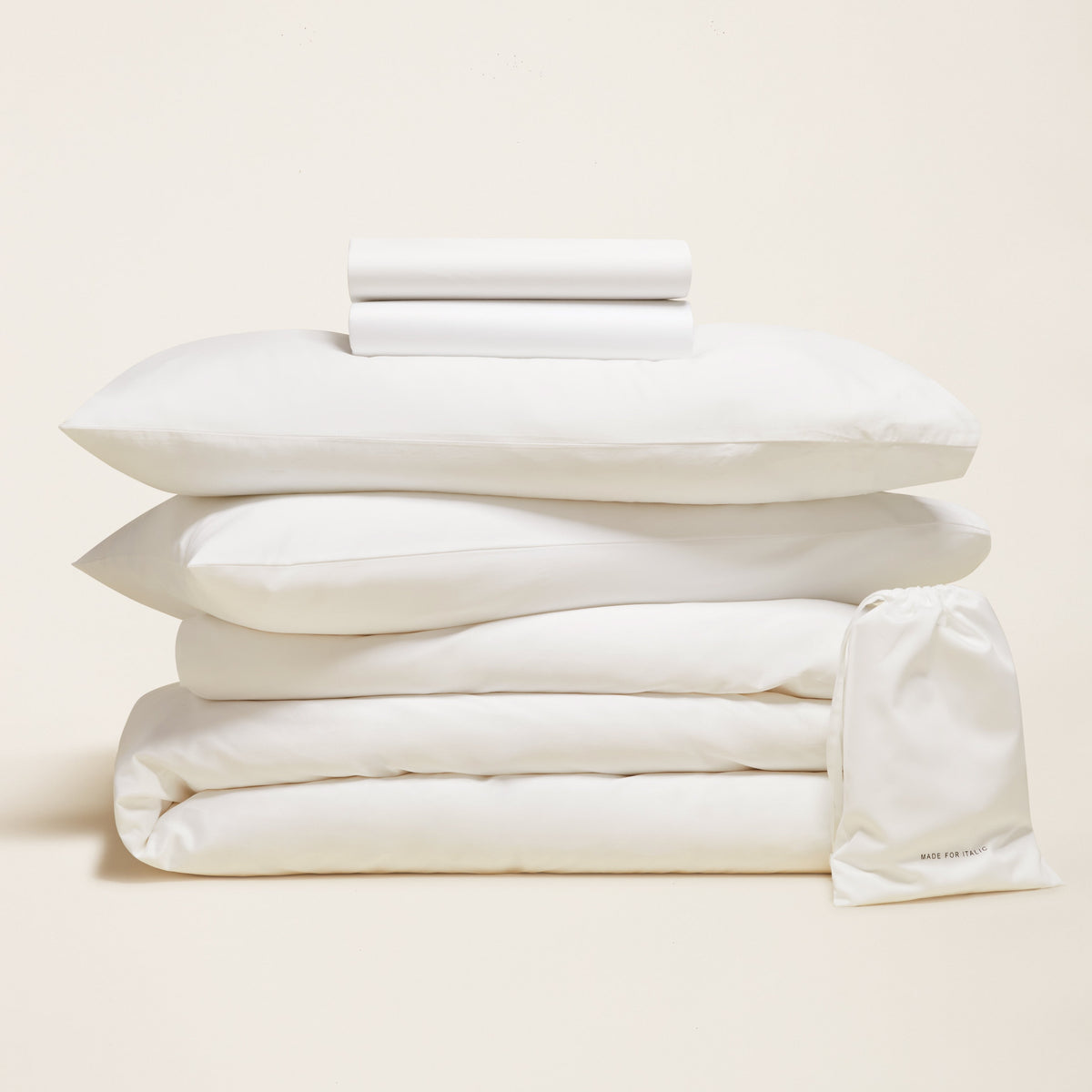 Folded white bedding bundle stacked centrally on a cream background beside a branded storage bag