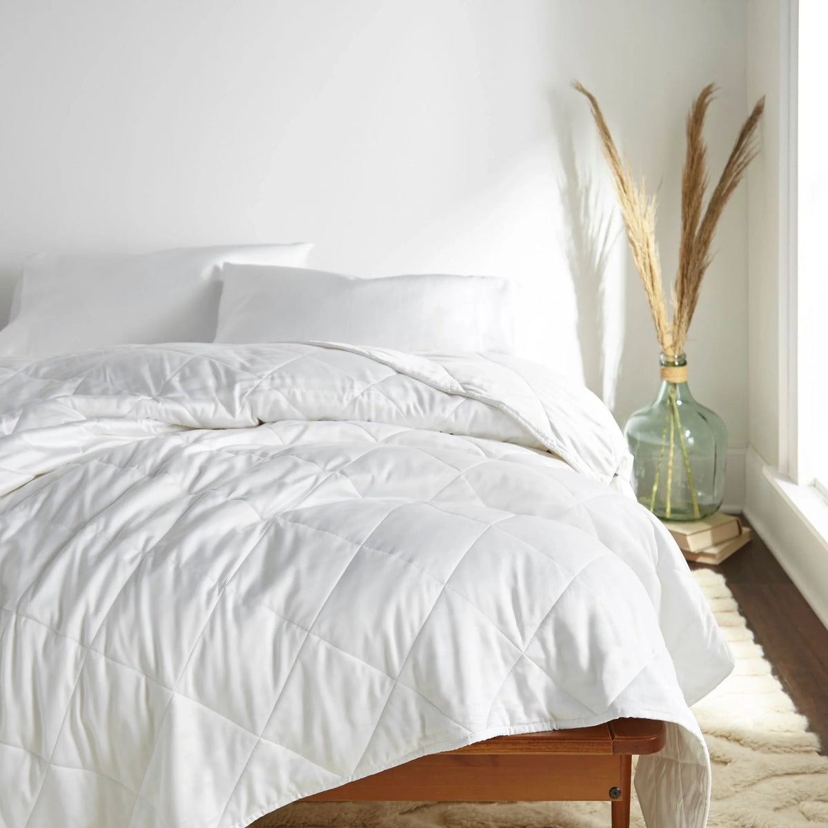 White diamond-quilted duvet draped over a wooden bed frame in a bright minimalist room with white pillows and dried pampas