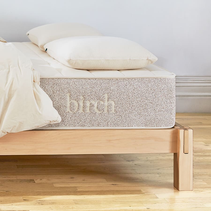 Close-up of cream organic cotton mattress corner with speckled grey side panel and 'birch' embroidery on light wood frame