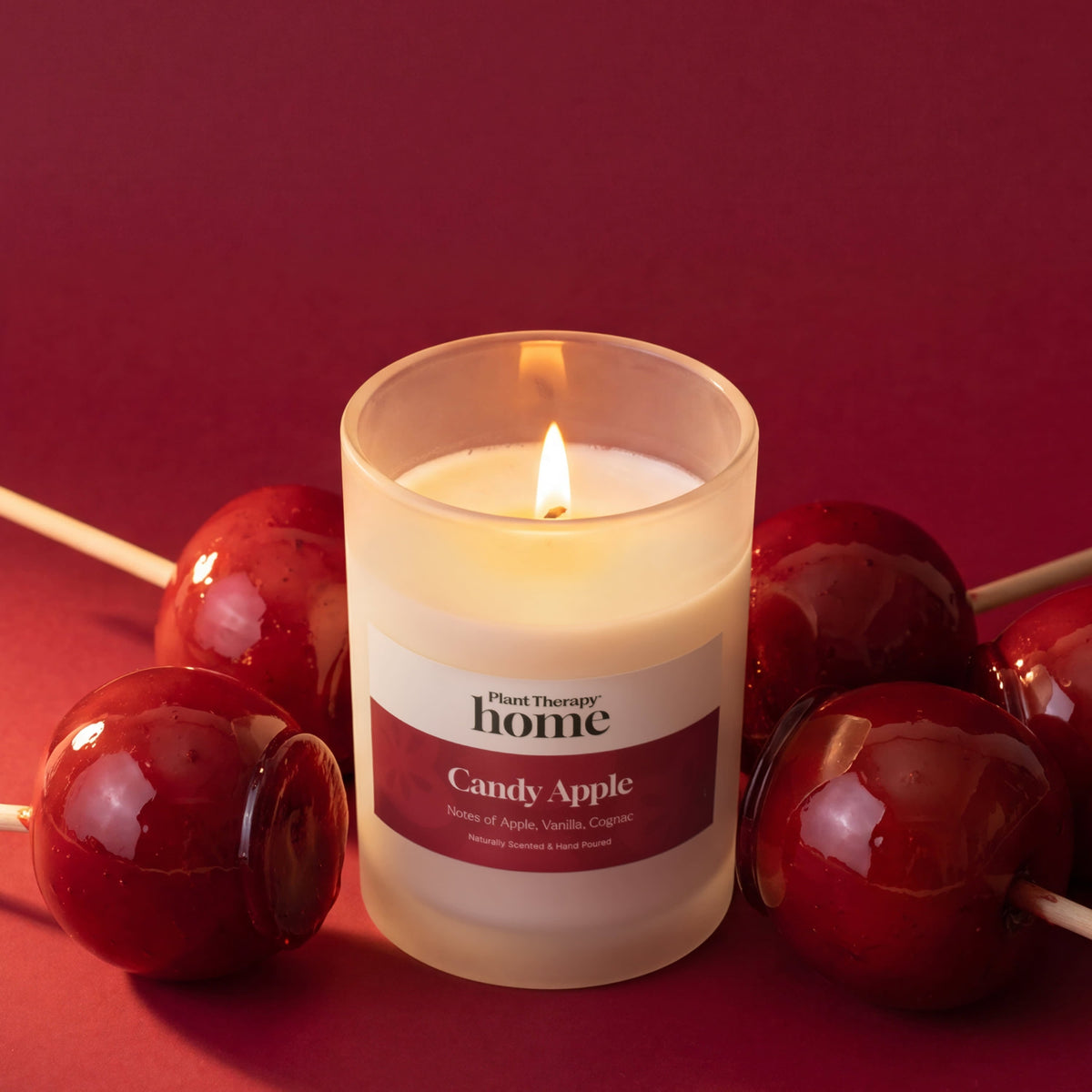 Lit candle in frosted jar surrounded by glossy red candy apples on sticks against deep red background