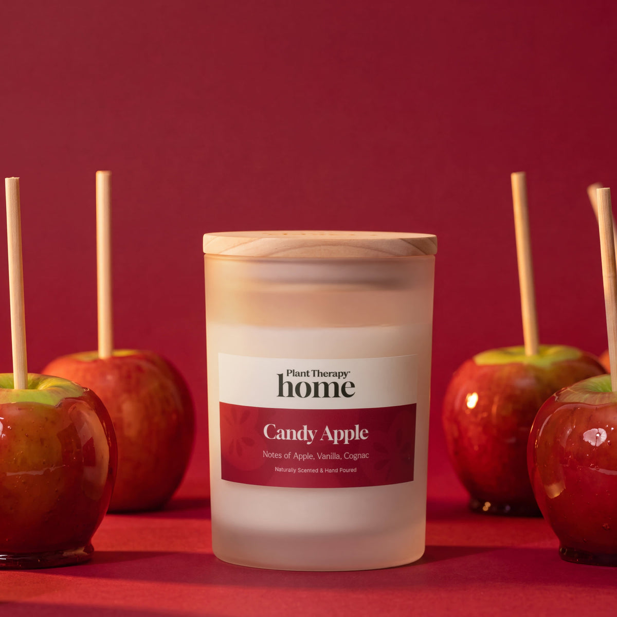Candle in frosted jar positioned among caramel coated apples on sticks against monochromatic red backdrop