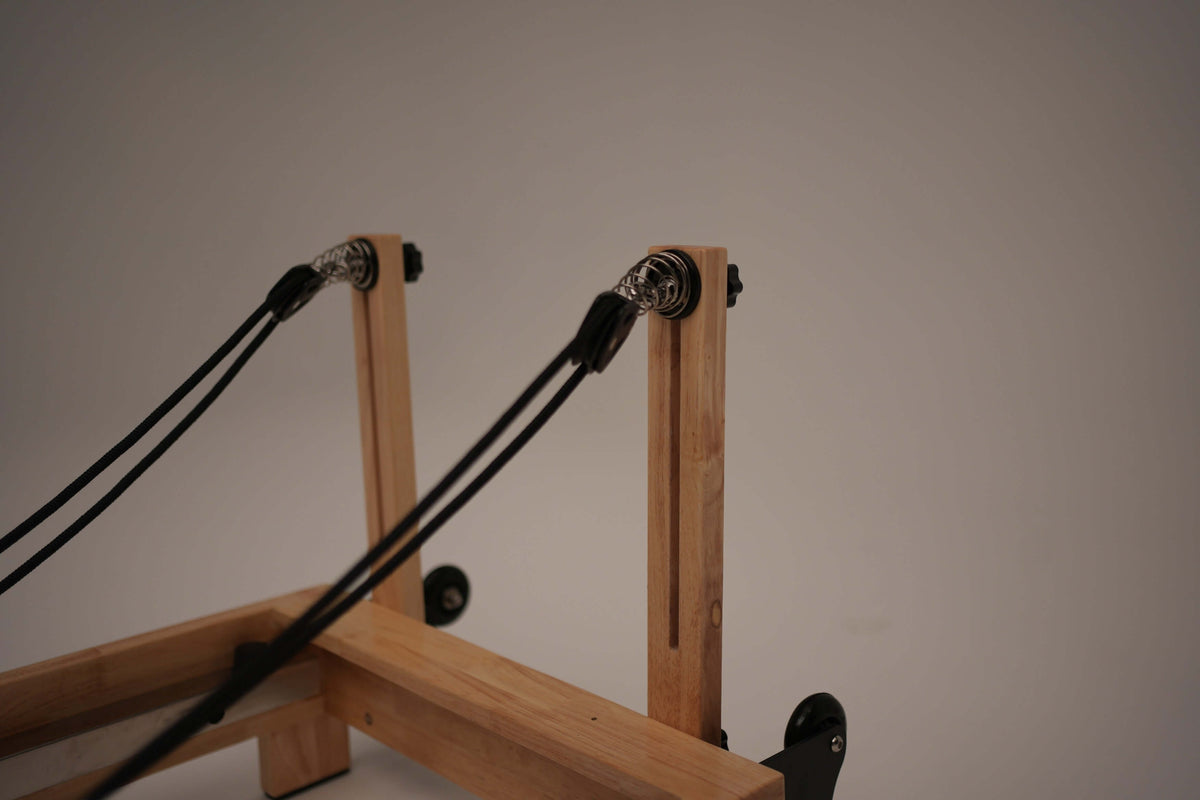 Rear perspective of reformer frame displaying vertical wooden posts, silver coil springs, and black ropes extending from