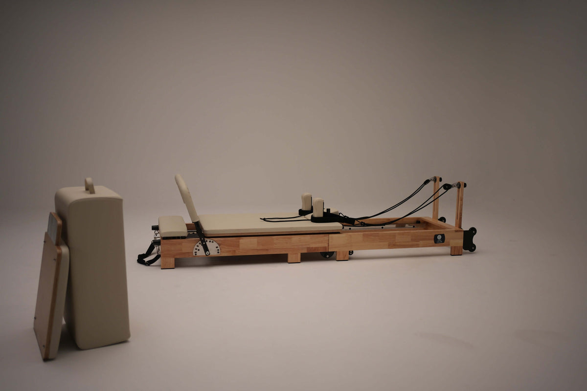 Light wood foldable Pilates reformer extended with beige upholstery, jumpboard accessory on left against grey background