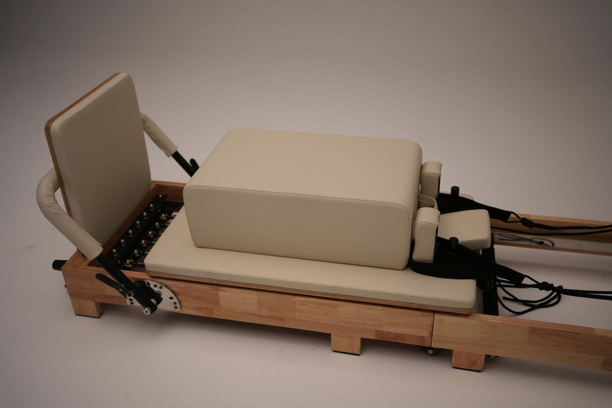 Studio shot of beige and light wood reformer with padded carriage, rectangular box attachment, vertical backrest, and visible