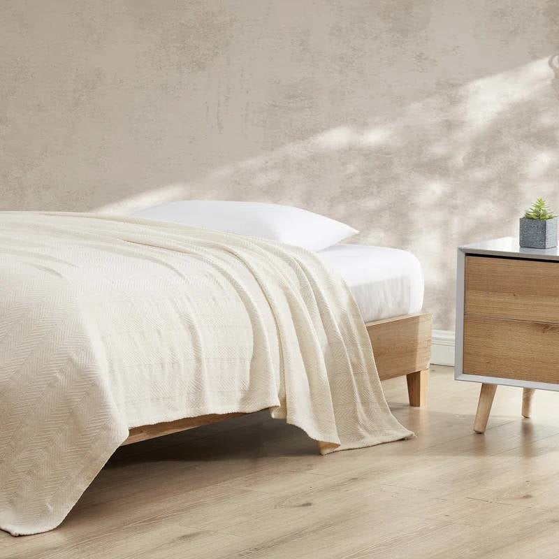Cream bone knitted cotton blanket draped over a wooden bed corner with white linens and beige wall