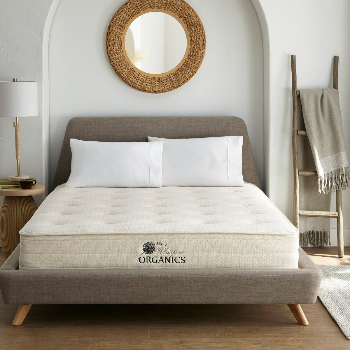 White Whisper Organics mattress on grey upholstered bed frame with wooden legs, two white pillows, and dandelion logo patch