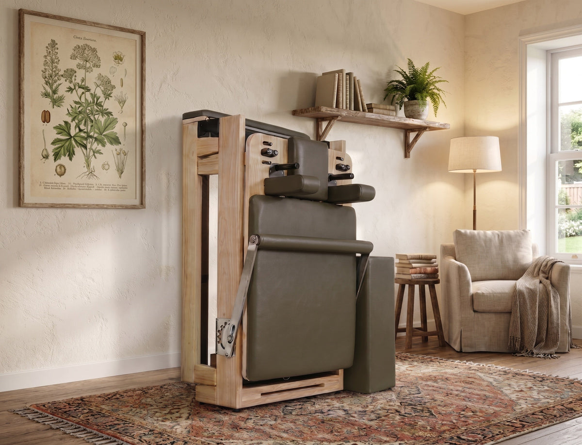 Folded olive green and light oak wood reformer standing vertically in a rustic living room with botanical art.