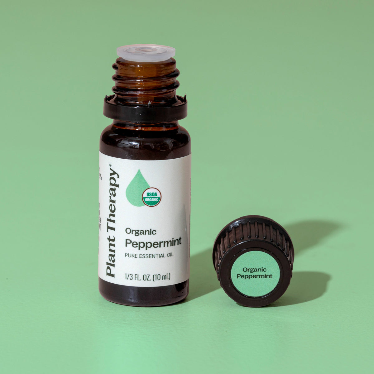 Open amber bottle of peppermint oil next to black cap on mint green background