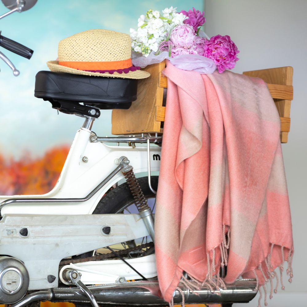 Cascading faded coral alpaca throw blanket draped over white vintage moped side next to straw hat and flowers