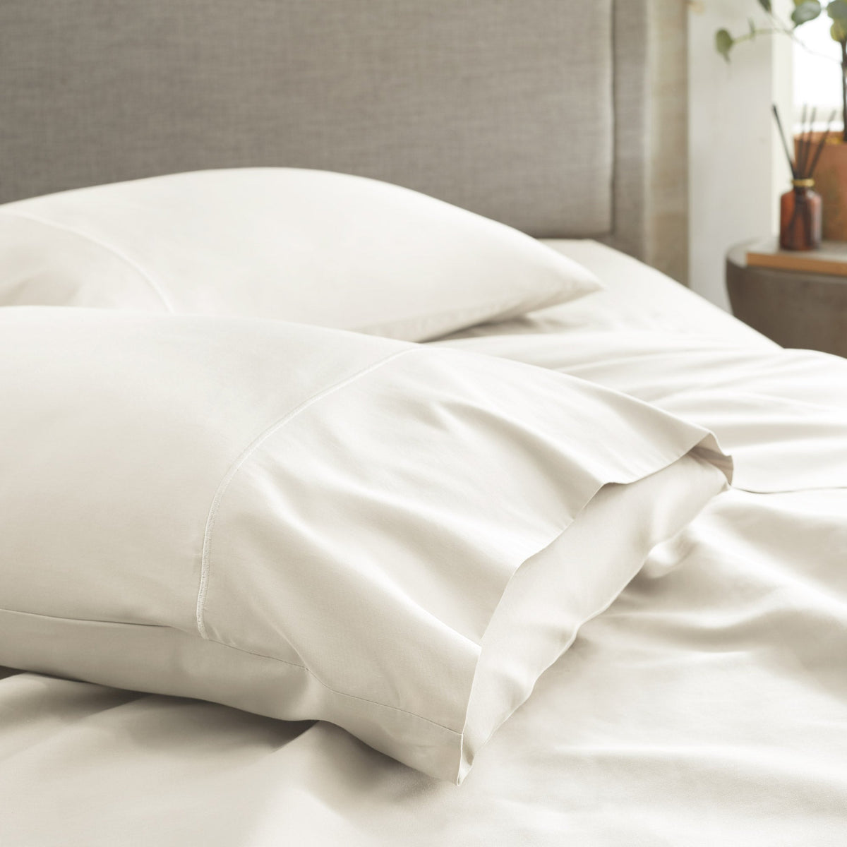 Off-white sateen pillowcases draped softly on a bed near a grey upholstered headboard