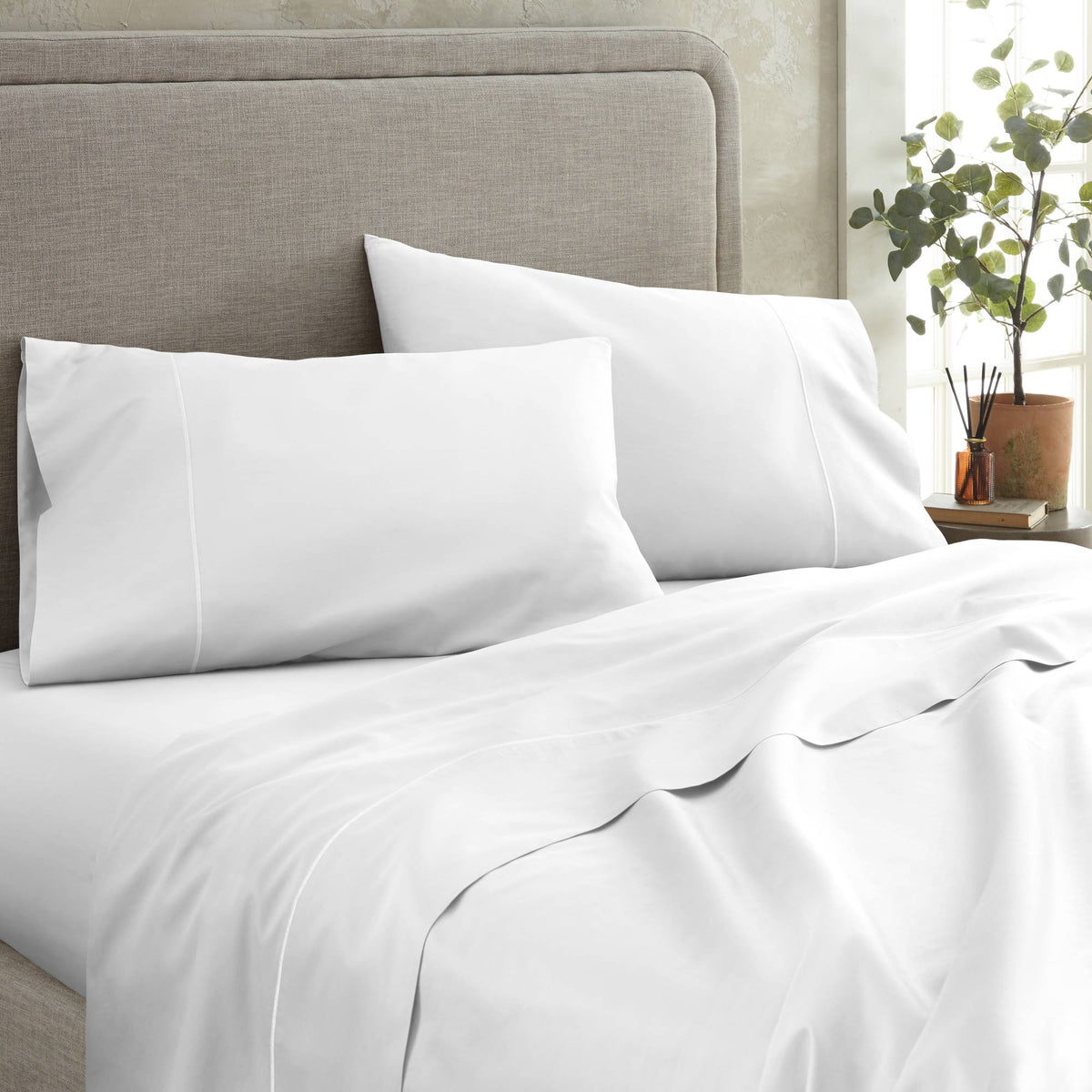 Crisp white pillowcases and fitted sheets with subtle sheen and elegant piped edge detail against grey headboard