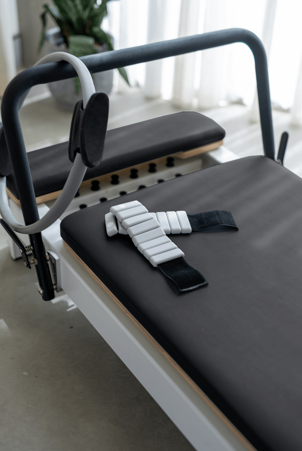 White reformer frame with black upholstery showing grey resistance ring and white ankle strap near plant background