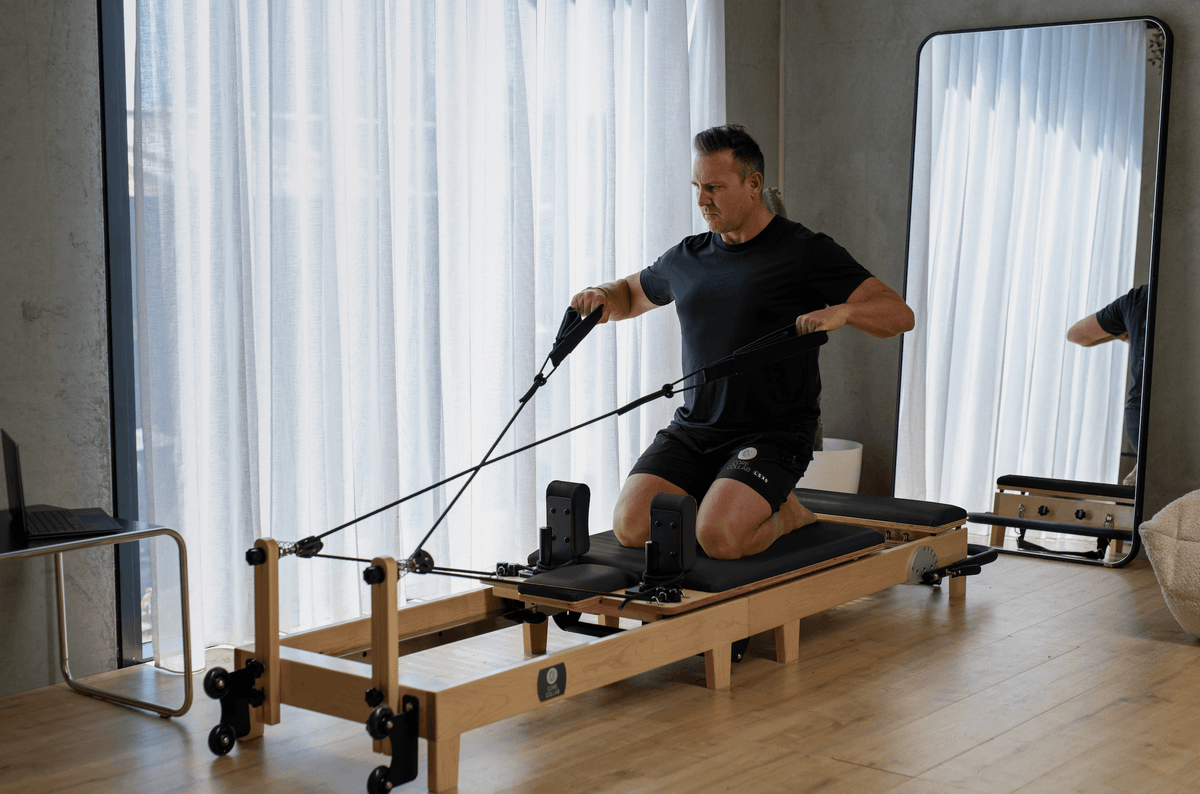 Instructor pulling black cable handles while kneeling on reformer carriage in bright studio with mirrors