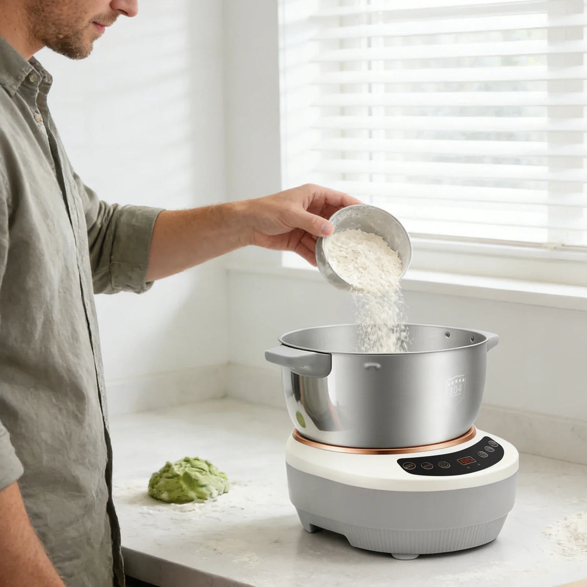 Man pouring flour from a measuring cup into the open stainless steel bowl of a silver dough mixer, with green dough on the