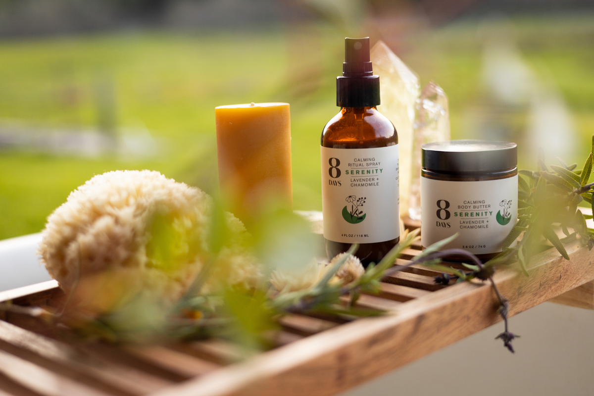 Eye-level shot of amber spray bottle and white body butter jar on wooden caddy with yellow candle, sponge, rosemary, and