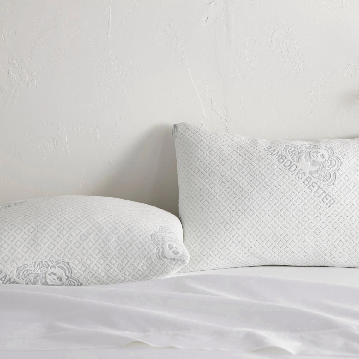 Two white diamond-quilted pillows with grey 'BAMBOO IS BETTER' panda logo on a bed