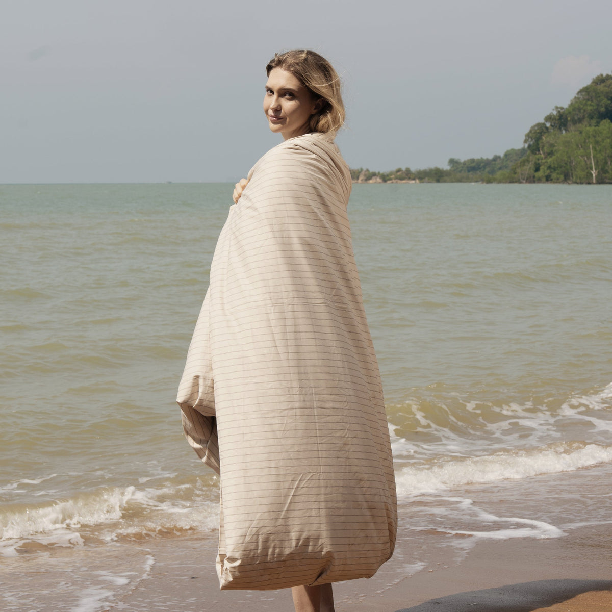 Woman wrapped in beige striped duvet cover on sandy beach near ocean waves