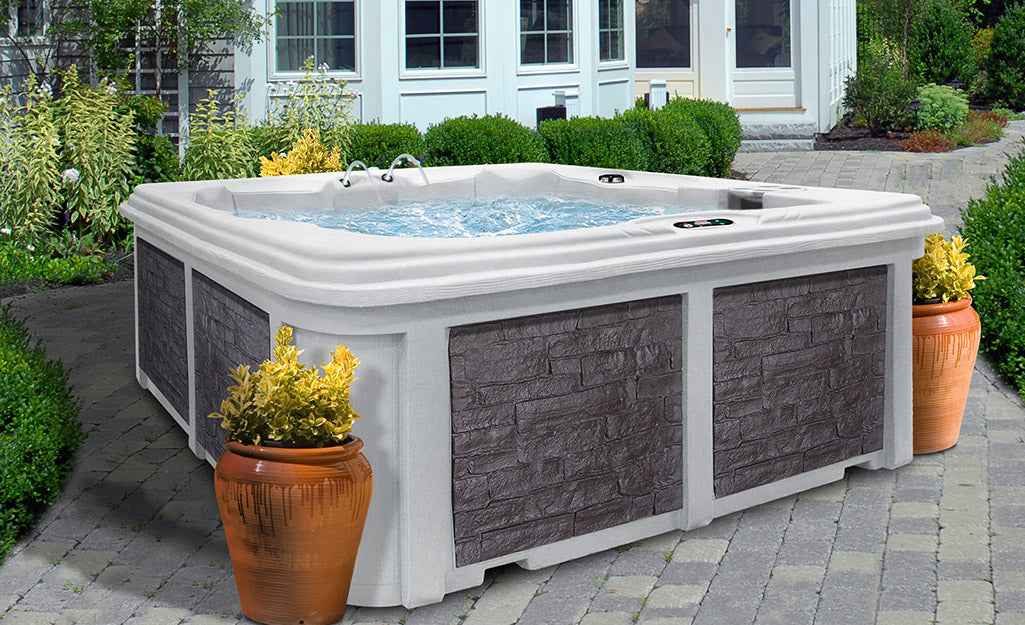 White acrylic hot tub with grey faux-stone panels on a patio, surrounded by terracotta planters and active water jets
