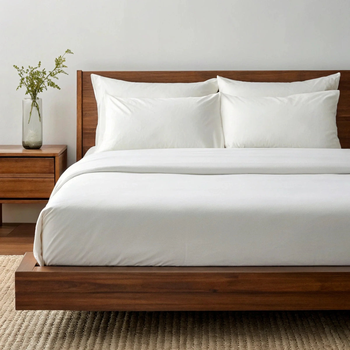 Angled perspective of white fitted sheet draping over dark walnut bed frame corner next to glass vase on nightstand