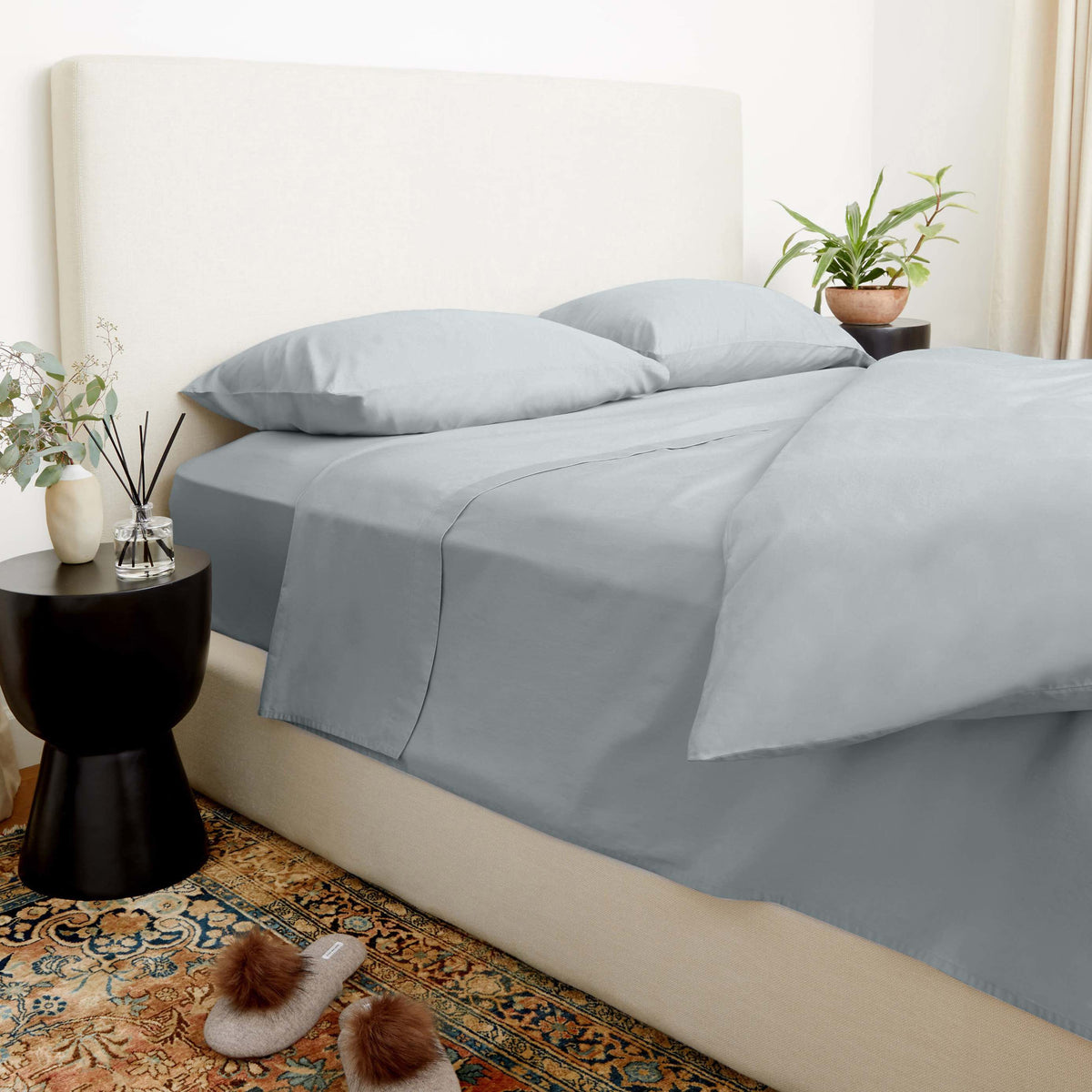 Solid slate-blue sateen bedding set on a beige upholstered frame styled with a reed diffuser and plant