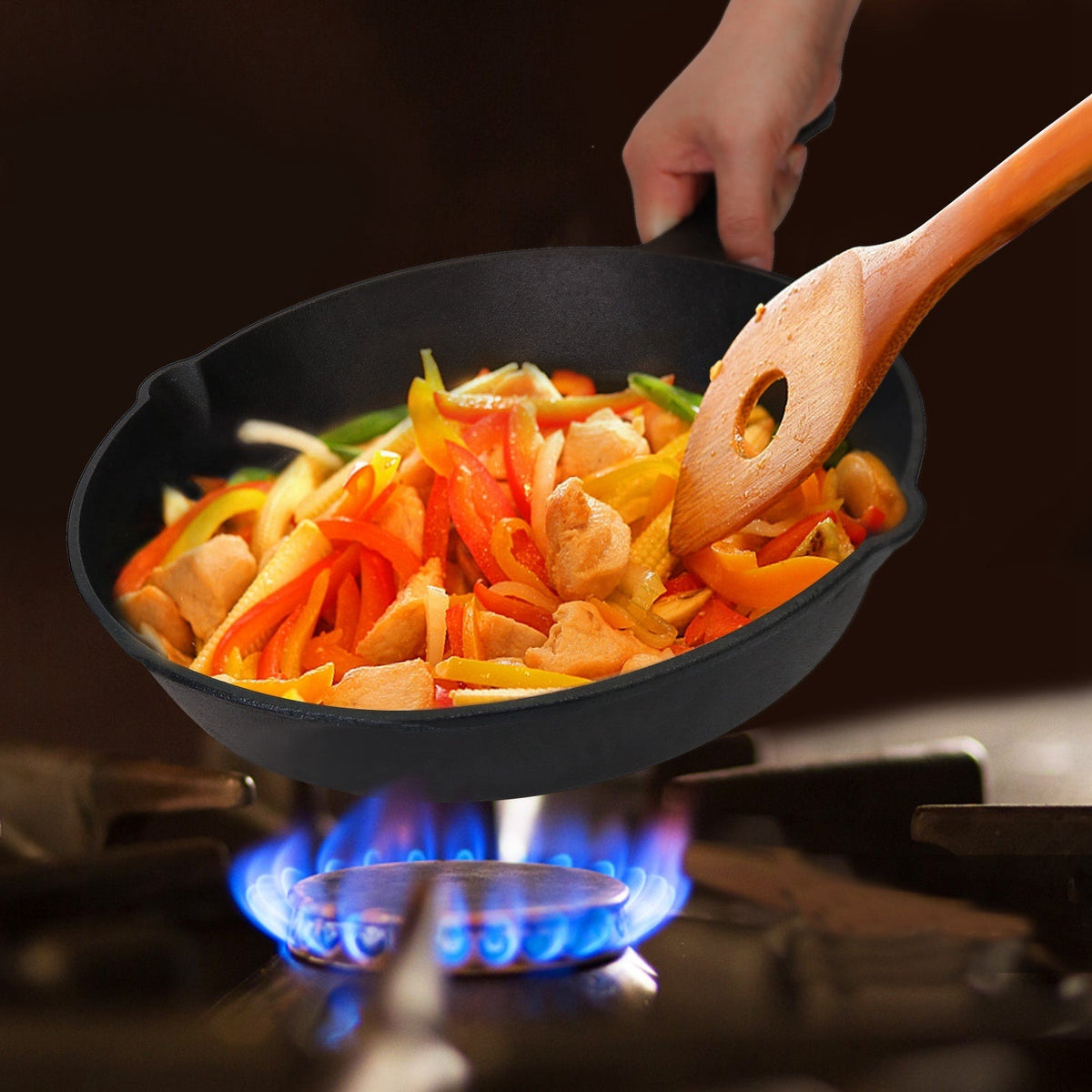 Hand stirring chicken and vegetables in black cast iron skillet over gas burner using wooden spatula with hole