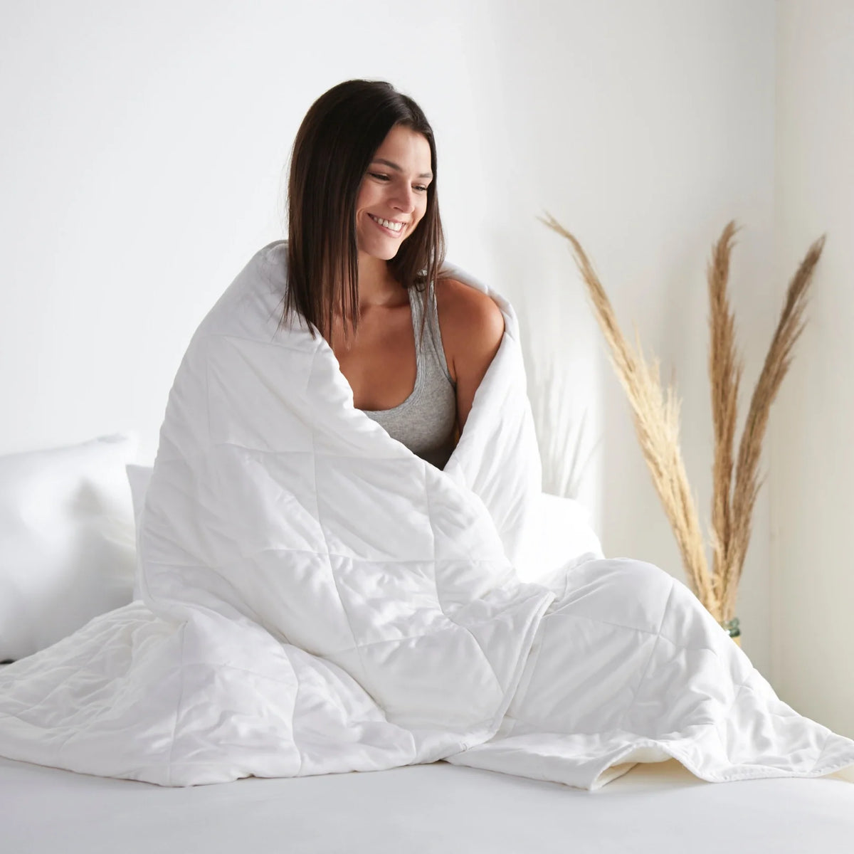 Smiling woman sitting on a bed wrapped in a white quilted duvet, showcasing the soft texture in a sunlit room with floral