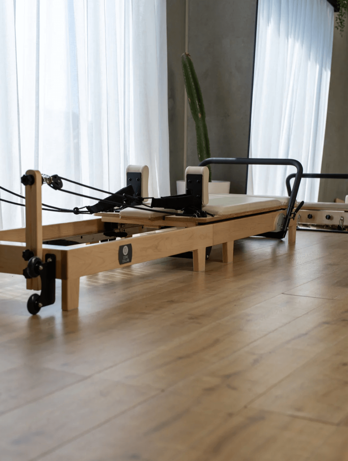 Low-angle perspective of reformer on light wood flooring highlighting sturdy legs, sliding carriage, black footbar, and logo