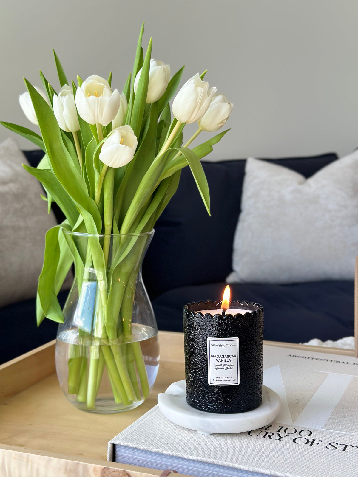 Lit black textured candle on white marble coaster beside fresh white tulips in glass vase on wooden tray