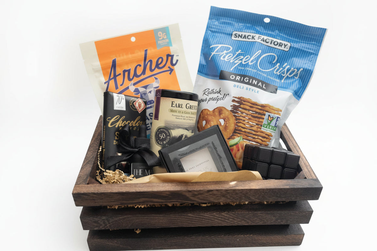 Unwrapped dark wooden crate displaying beef jerky, pretzel crisps, dark chocolate, tea, and square black soap bar