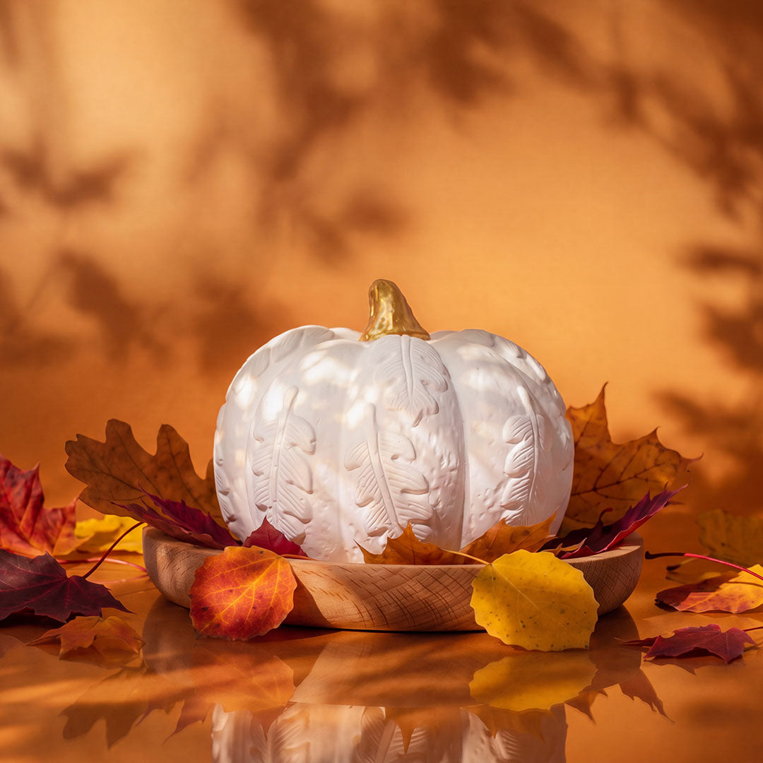 White plaster pumpkin diffuser with gold stem on beech wood tray surrounded by autumn maple leaves on orange background