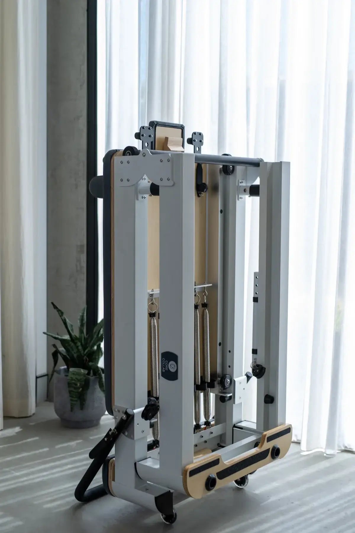 Folded white steel reformer with exposed springs, wooden accents, and wheels standing near sheer curtains