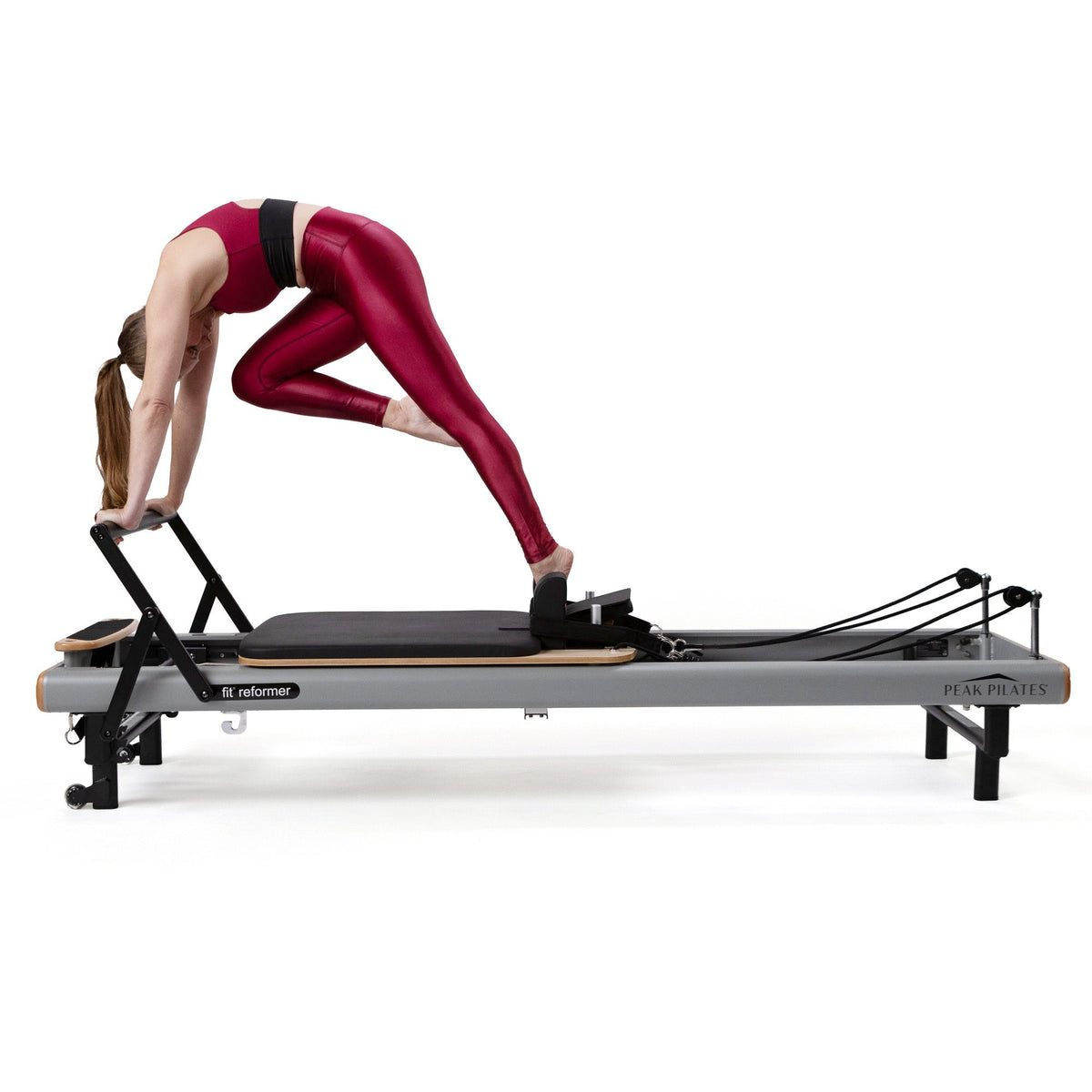 Female athlete in red activewear performing arched pose on grey fit Reformer, gripping overhead bar while demonstrating