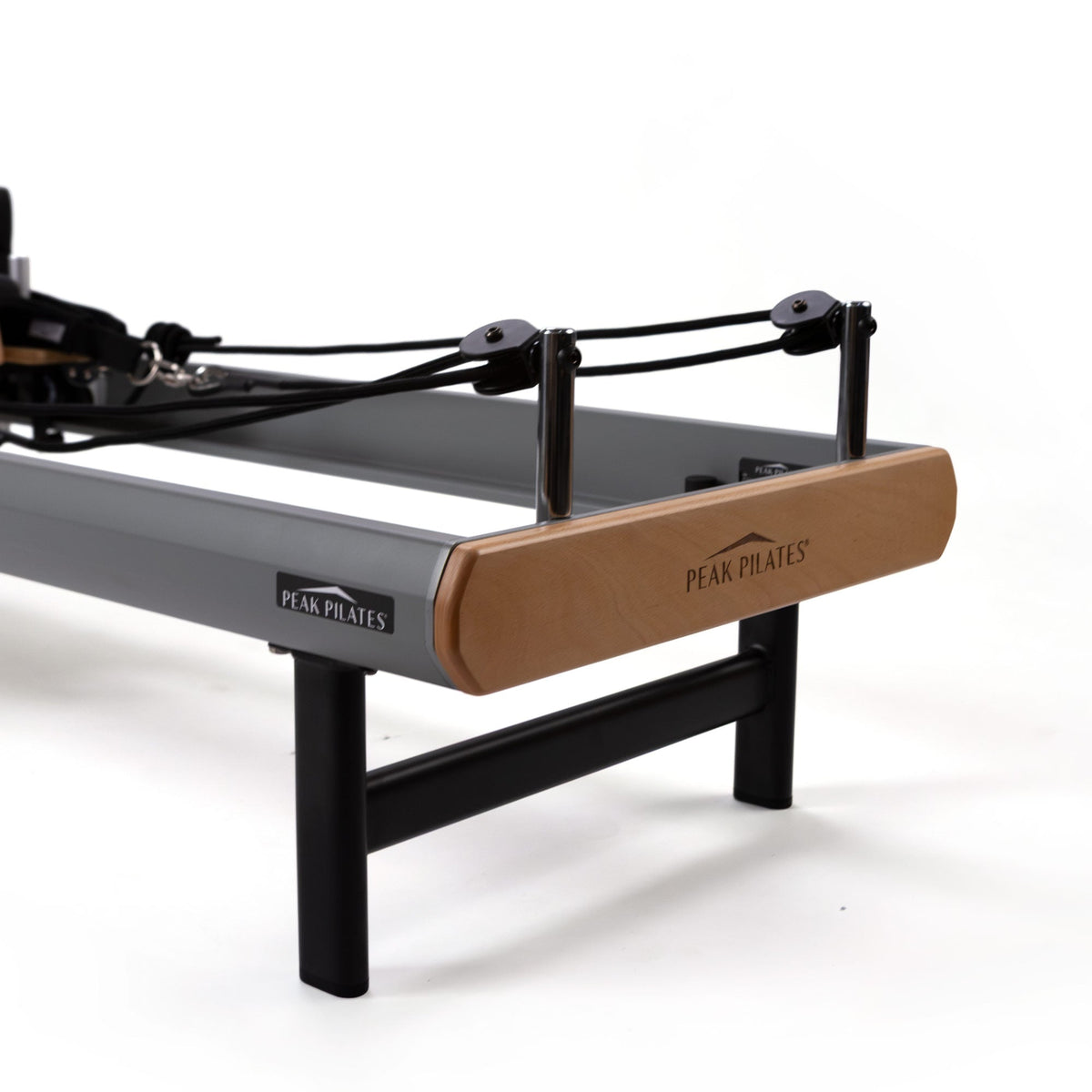 Close-up of light wood cap with logo, chrome uprights, black cords, and sturdy legs at footbar end of reformer