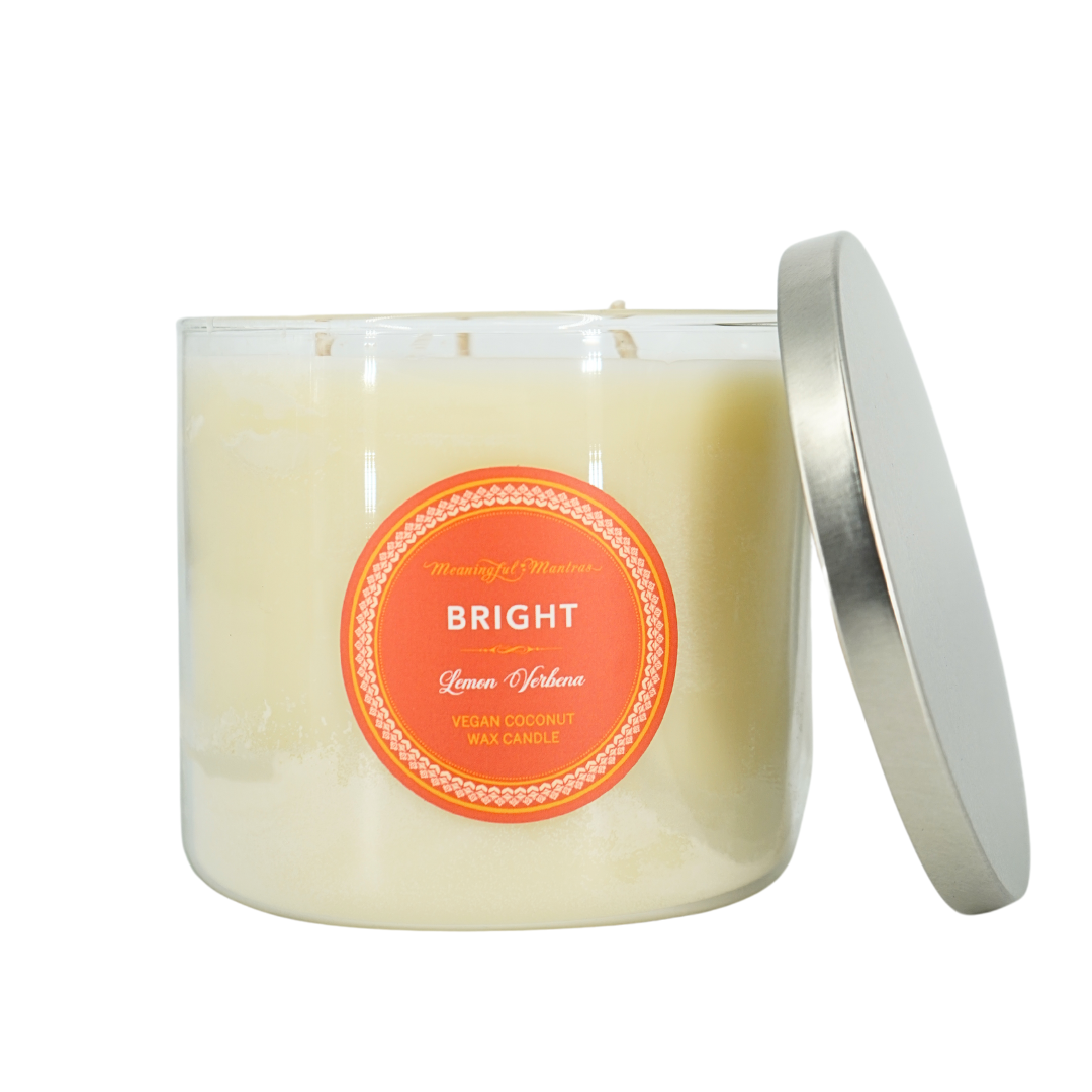 Clear three-wick glass candle with orange sticker and silver metal lid leaning against the side