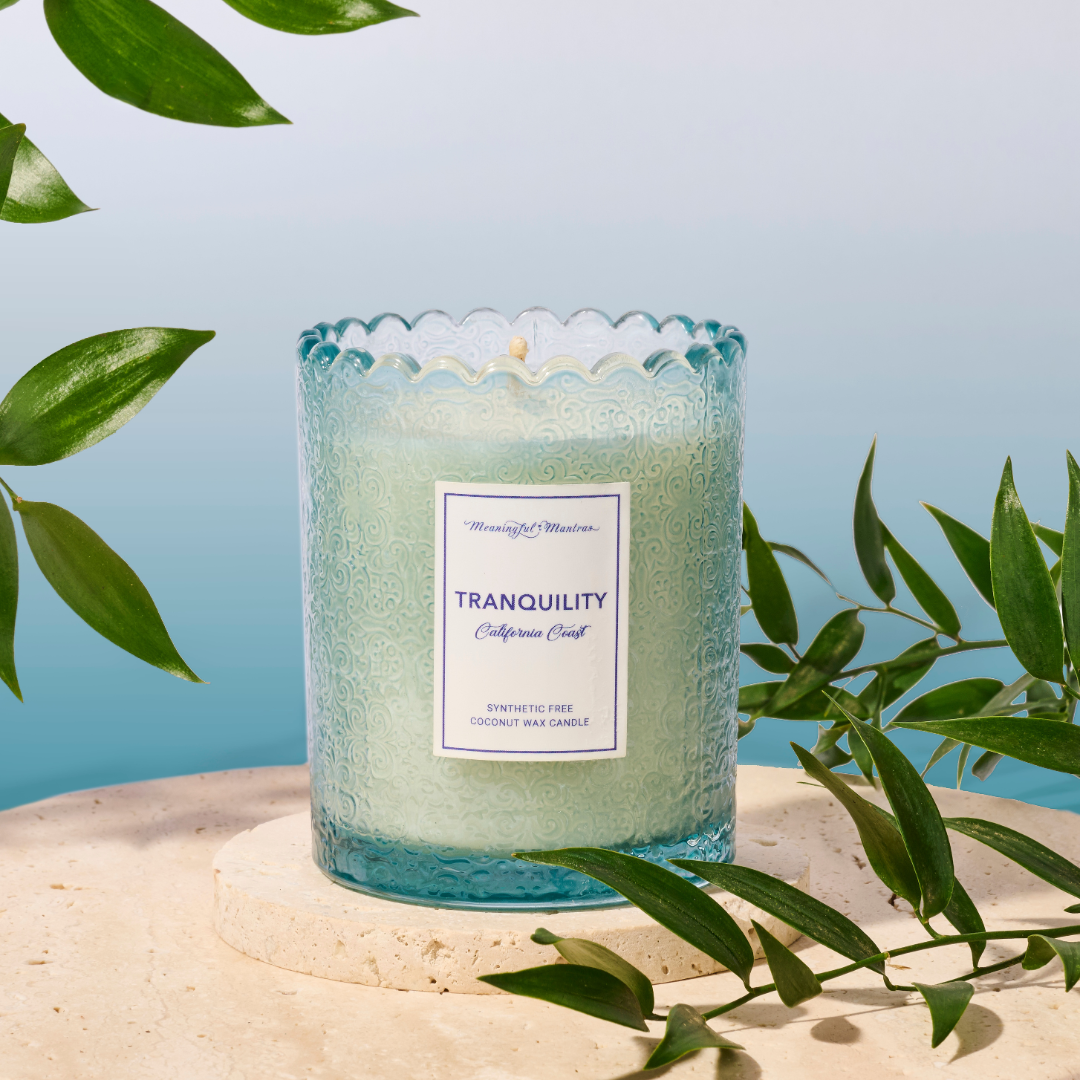 Blue textured coconut wax candle on beige stone coaster surrounded by green leaves against blue background