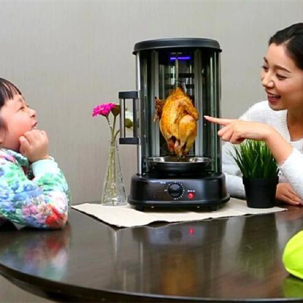 Black electric rotisserie grill with glass cylinder containing a rotating golden-brown chicken, viewed by a woman and child