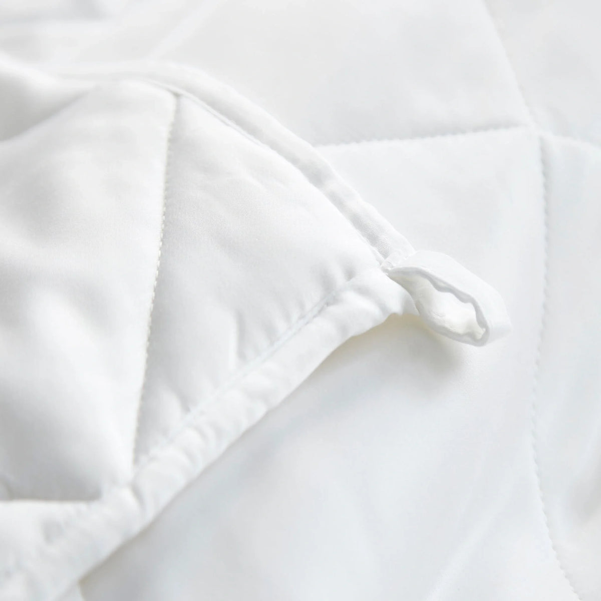 Corner detail of a white comforter featuring a small fabric tie loop for securing to a cover, with clean hemmed edges and