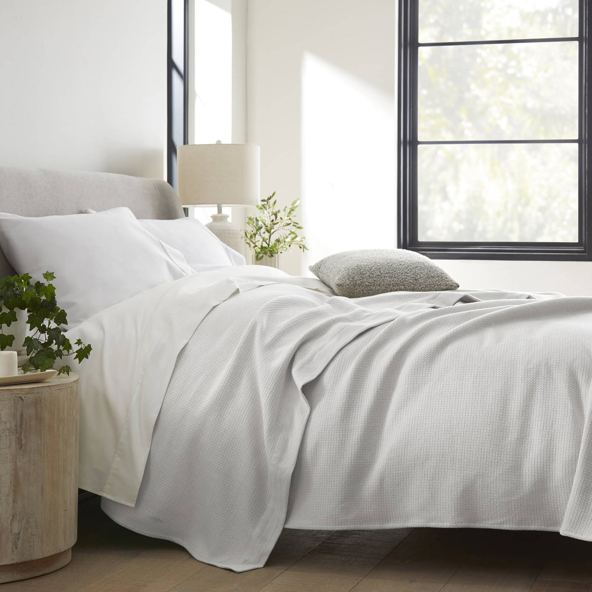 Light grey woven blanket draped on bed with white linens, plant, and sunlight in bedroom