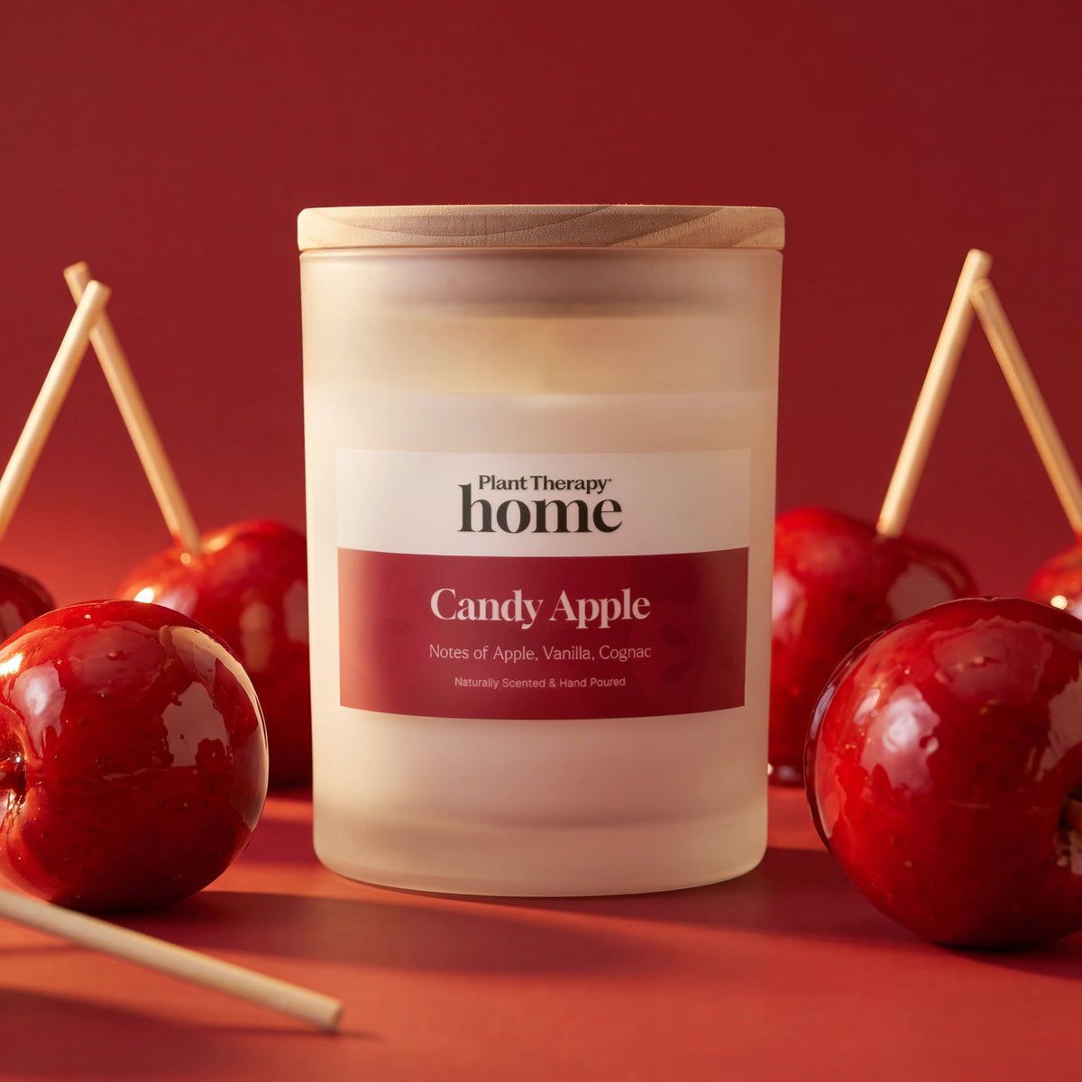 Frosted white candle jar with wooden lid centered on red surface surrounded by red candy apples