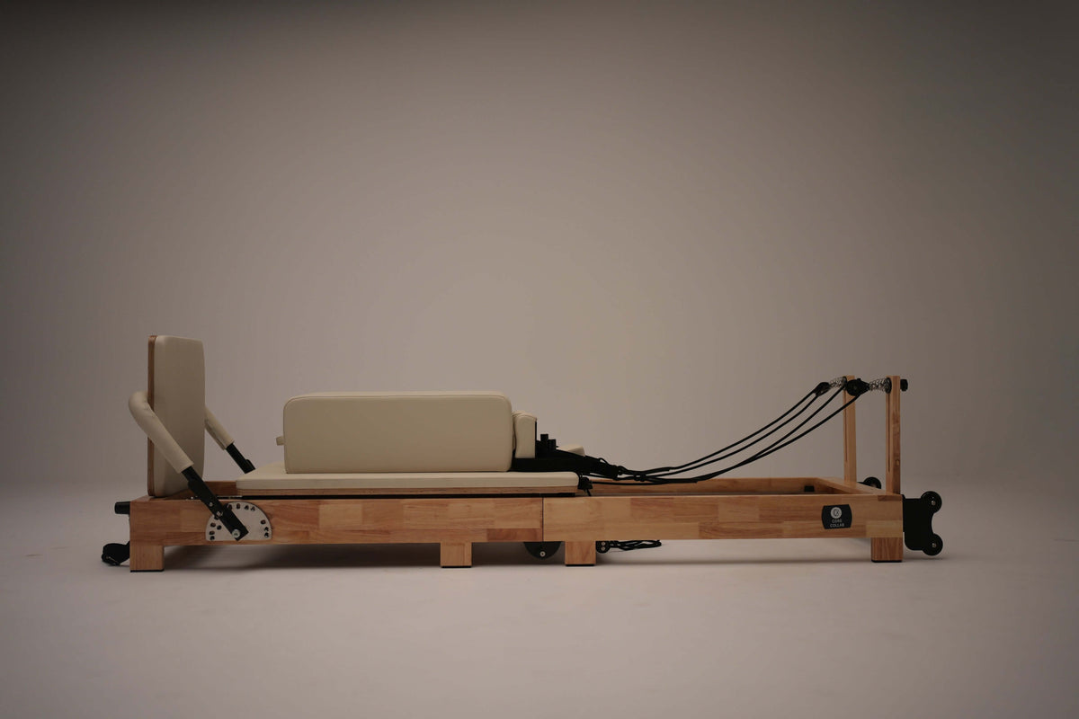 Side profile of light natural wood reformer with beige upholstered carriage, black suspension ropes, and spring tension dial