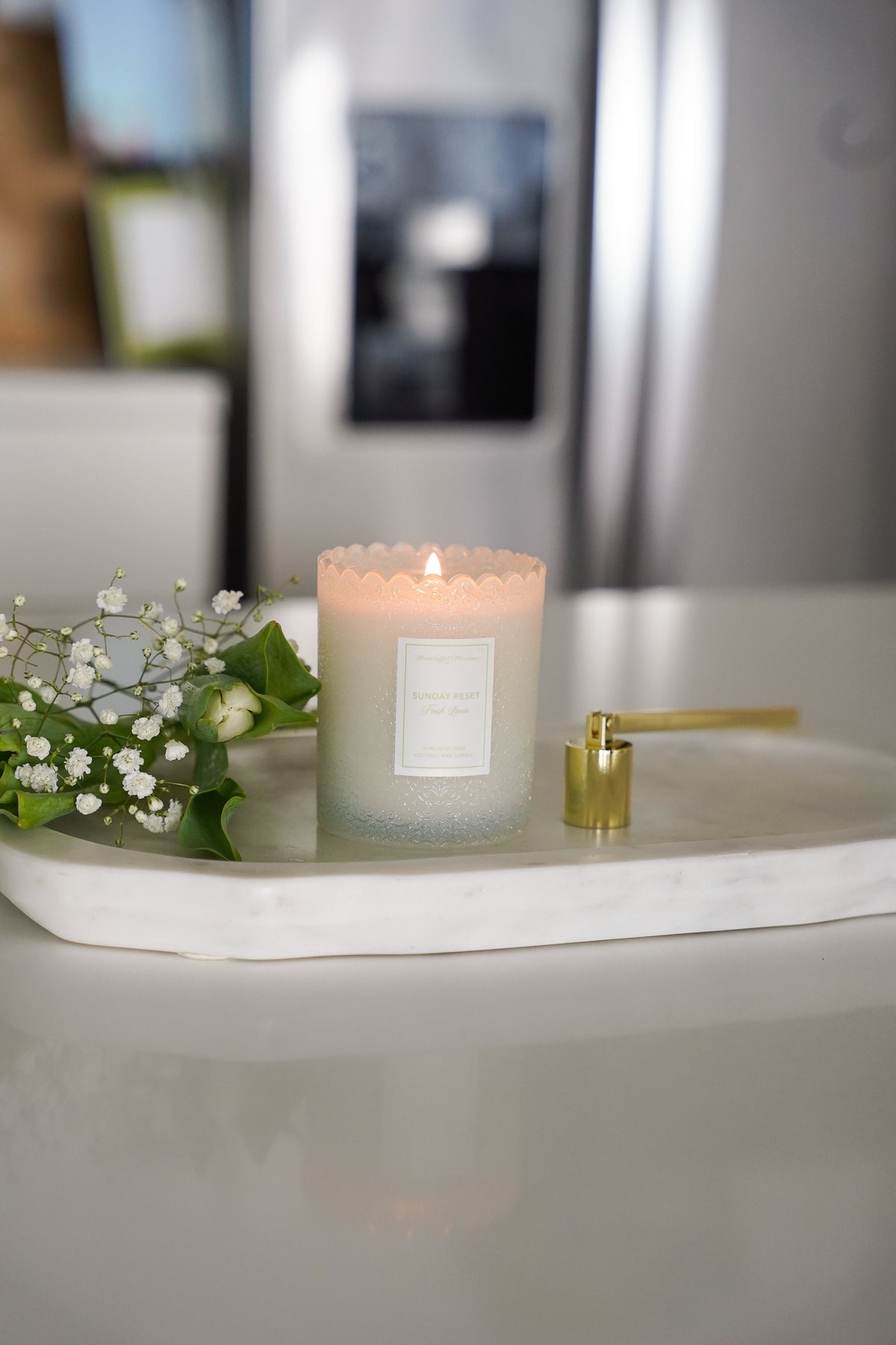 Lit coconut wax candle glowing warmly on marble tray next to white flowers in modern kitchen