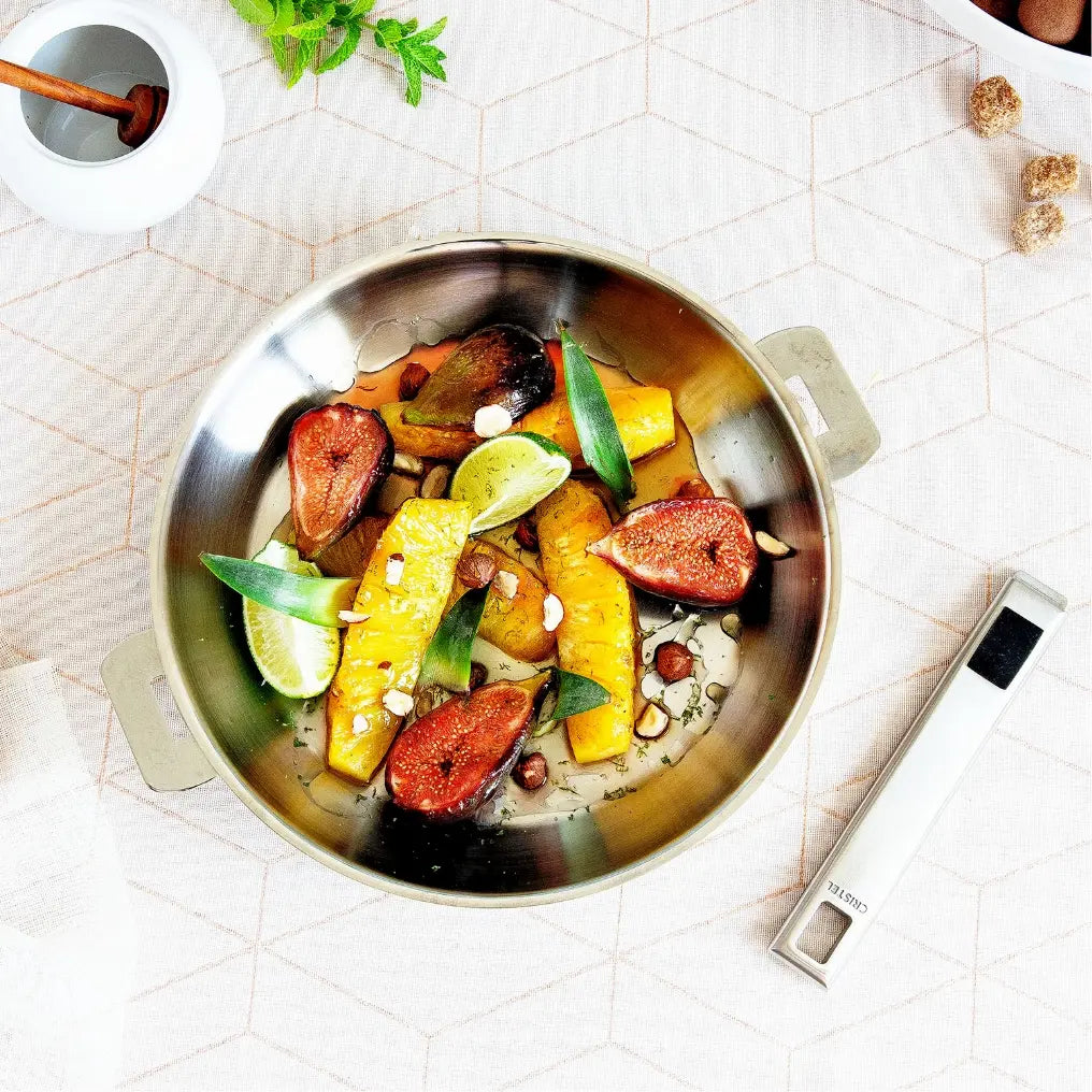 Stainless steel deep frying pan serving roasted figs and squash on a patterned tablecloth with detached handle nearby