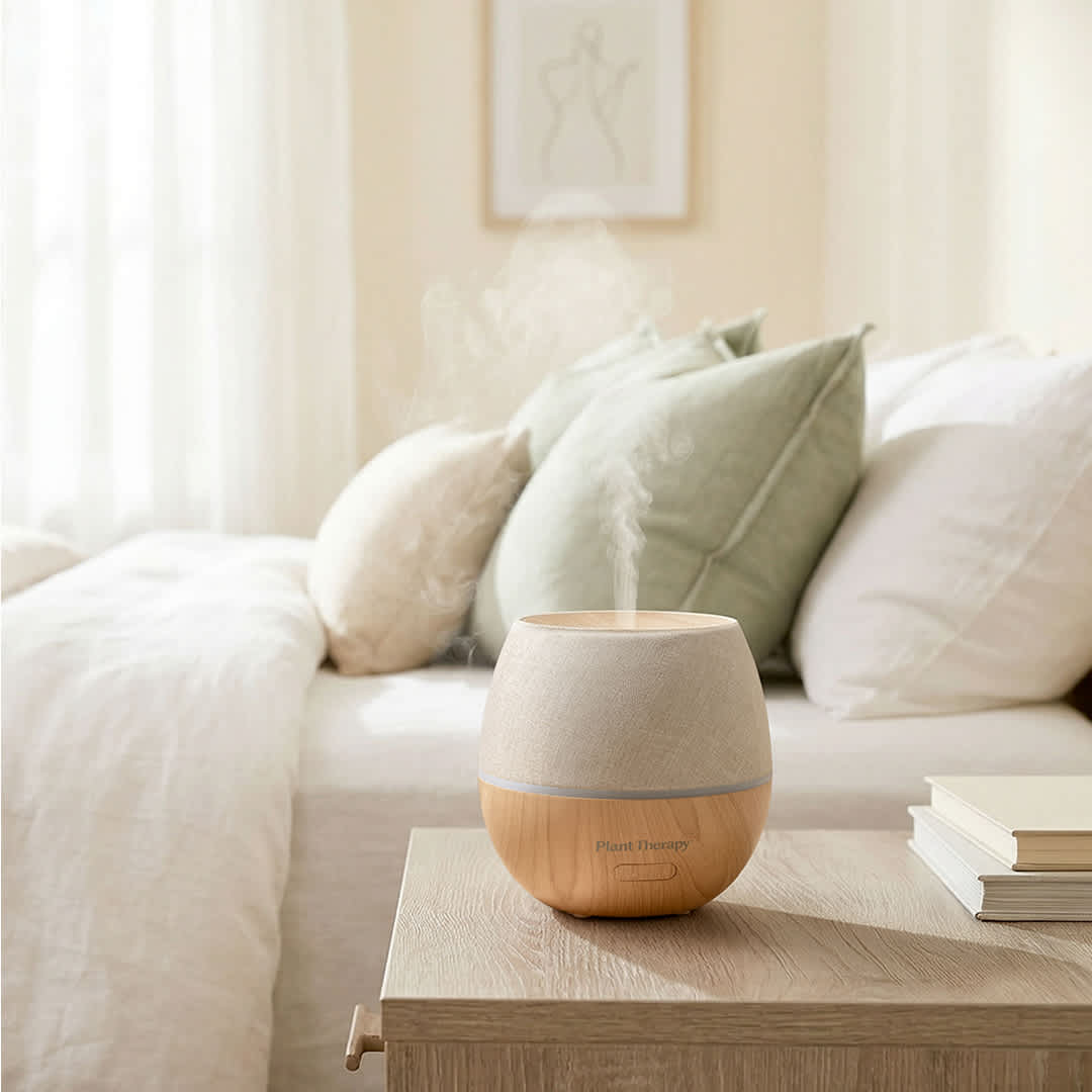 Diffuser emitting mist on bedroom nightstand with sage pillows and sheer curtains
