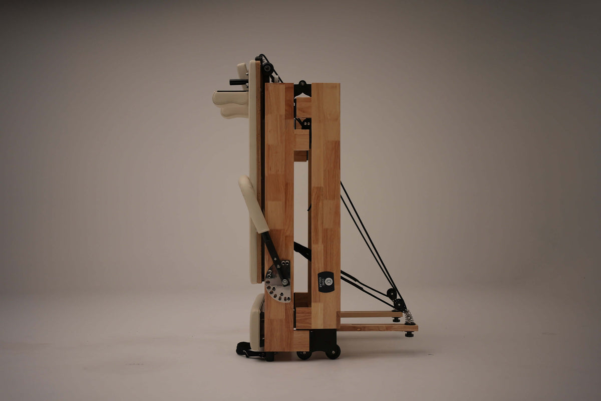 Vertical side view of folded light wood reformer revealing compact profile, internal springs, and base wheels for portability