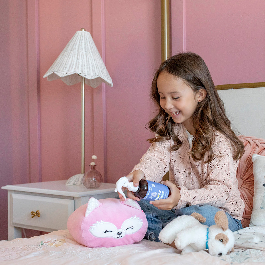 Young girl spraying pink fox pillow in bedroom while holding stuffed dog