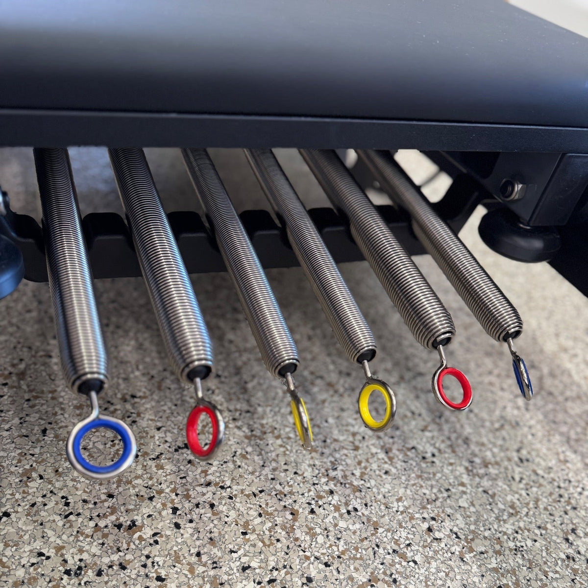 Close-up of six silver steel resistance springs with blue, red, and yellow color-coded rings hanging beneath the reformer