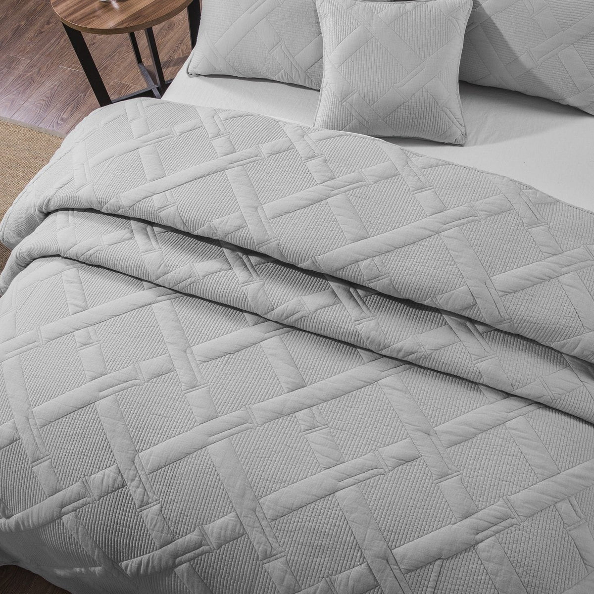 Overhead shot of light gray quilt draped over a bed corner with two square throw pillows, revealing detailed basket-weave