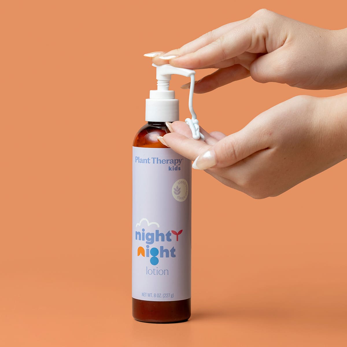 Hands pressing pump to dispense white lotion swirl onto finger against orange background