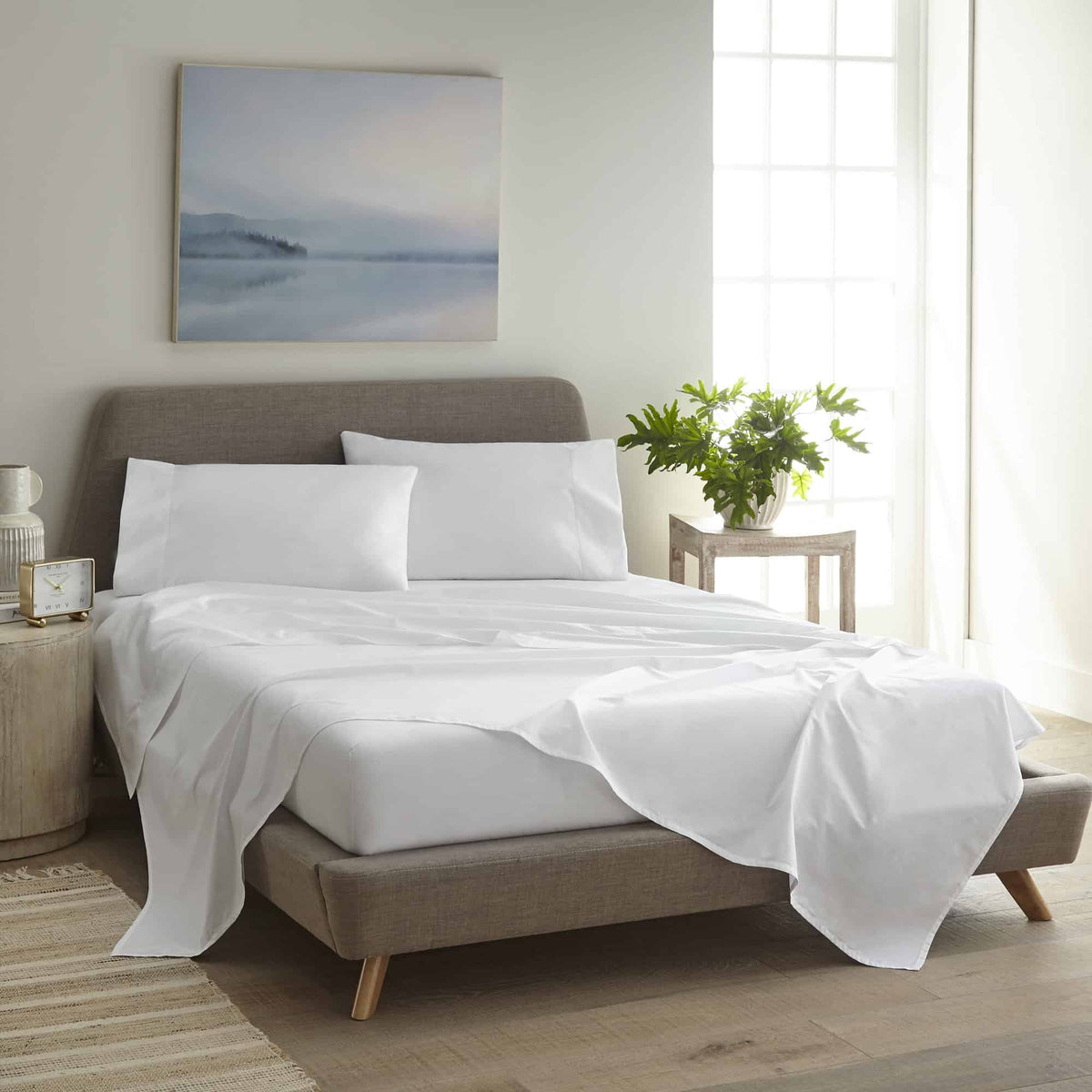 White sheet set draped over grey upholstered bed frame in bright bedroom with plant